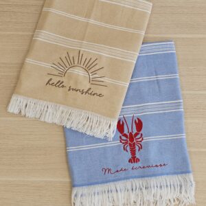 serviettes-fouta-personnalisees-broderies.jpg Deux serviettes fouta personnalisées avec broderies soleil et homard sur fond bois