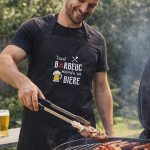 Homme portant un tablier noir personnalisé « Tout barbecue mérite sa bière » en train de cuisiner au barbecue