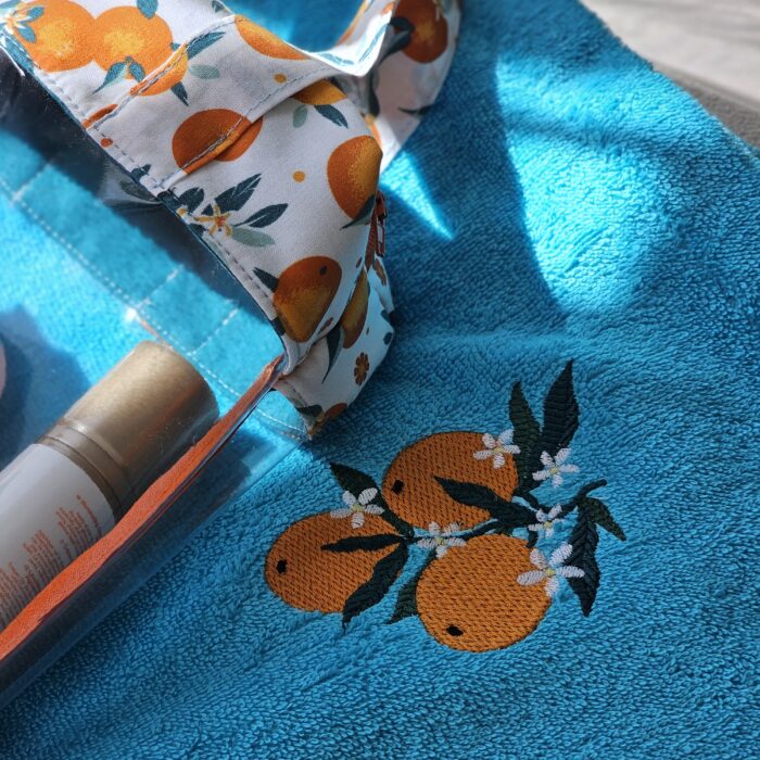 trousse-sac-maillot-orange-serviette-brodee.jpg Sac à maillot de bain motif oranges posé sur une serviette turquoise brodée avec des oranges